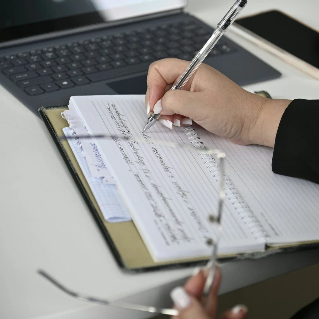 A person writing on a notebook with a laptop in the background